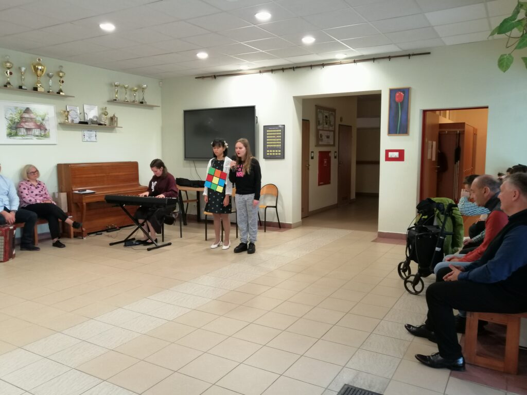 Zdjęcie przedstawia uczennice podczas występu artystycznego na szkolnym korytarzu z okazji, Dnia Edukacji Narodowej. Jedna dziewczyna stoi i śpiewa do mikrofonu, a druga gra na keyboardzie. W tle uczestnicy słuchają piosenki.