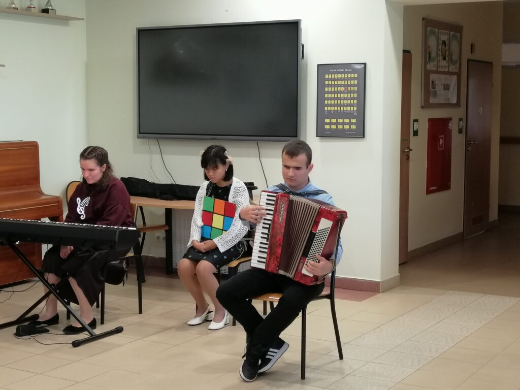 Zdjęcie ukazuje fragment występu artystycznego podczas uroczystości szkolnej, z udziałem trojga młodych ludzi. Uczeń po prawej gra na akordeonie, a dwie dziewczyny siedzą obok – jedna przy keyboardzie, a druga z dużą kolorową kostką. W tle widoczny jest duży ekran i pianino.