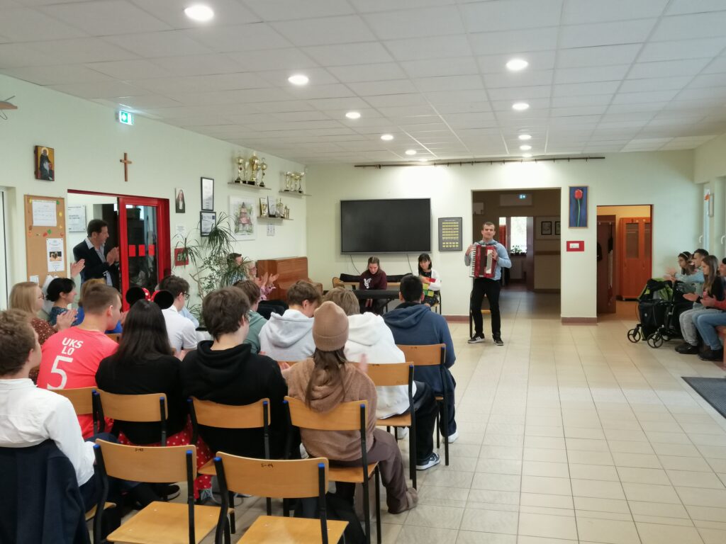 Zdjęcie ukazuje koniec występu ucznia na akordeonie i brawa uczestników. W tle widać uczniów i nauczycieli na korytarzu.