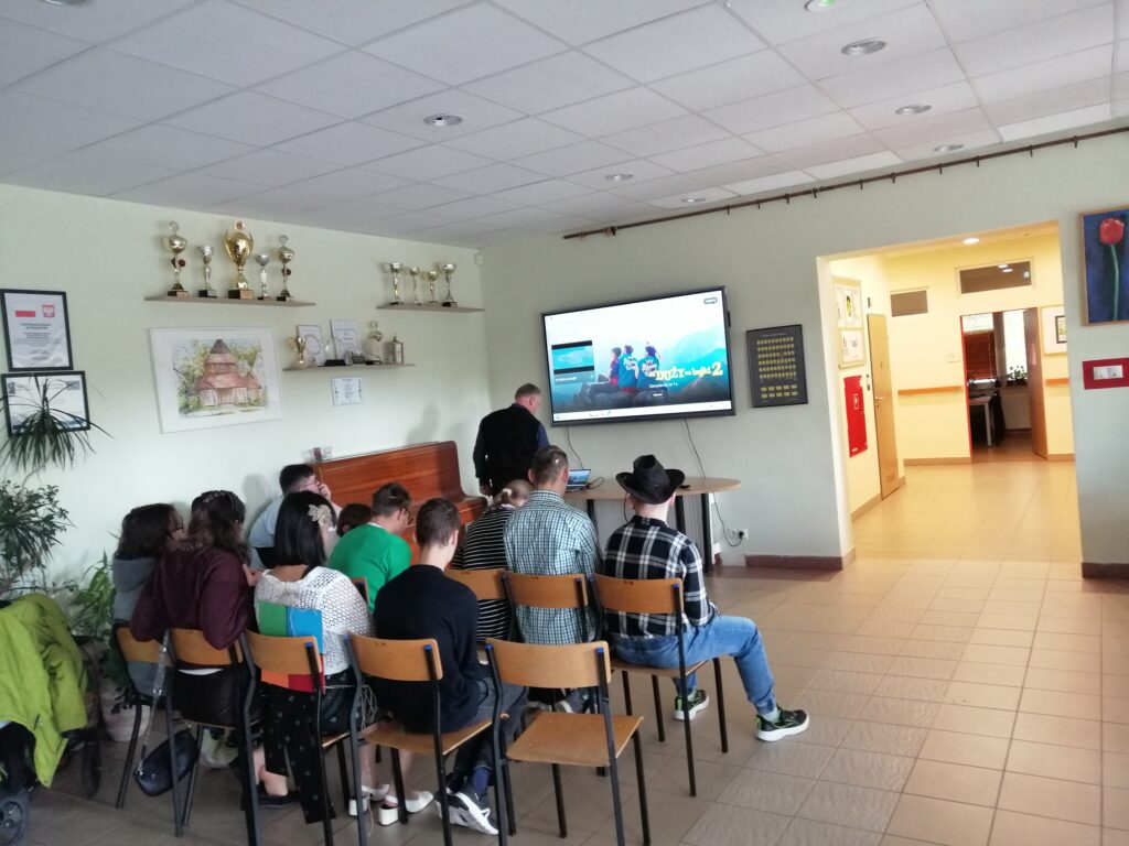 Zdjęcie przedstawia grupę uczniów siedzących na krzesłach, zwróconych plecami do obiektywu, obserwujących film na dużym telewizorze w szkolnym korytarzu.