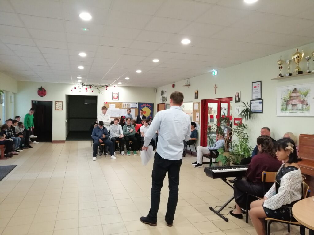 Zdjęcie przedstawia scenę z uroczystości szkolnej na korytarzu, gdzie widoczny jest nauczyciel odwrócony plecami, przemawiający do zgromadzonej młodzieży. Uczniowie pierwszej klasy stoją i słuchają przywitania wychowawcy.