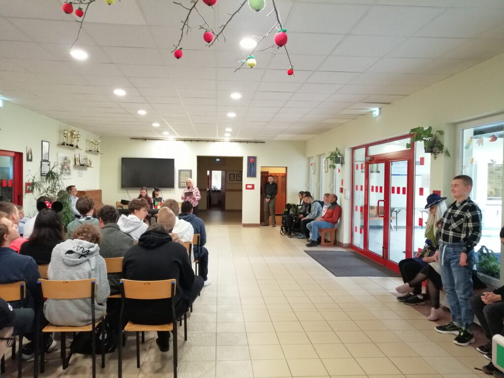 Zdjęcie ukazuje uczniów zgromadzonych na szkolnym korytarzu, siedzących w rzędach i zwróconych w kierunku głębi pomieszczenia. Wychowawczyni przemawia do uczniów wyczytując wychowanków swojej klasy.