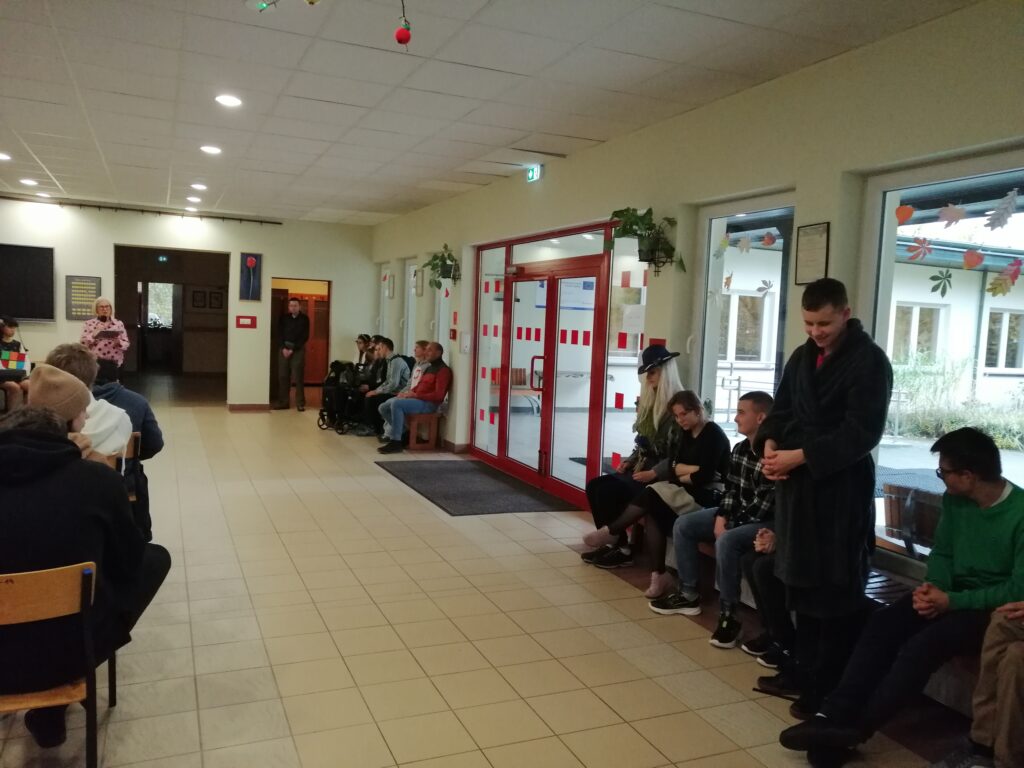 Zdjęcie ukazuje uczniów zgromadzonych na szkolnym korytarzu, siedzących w rzędach i zwróconych w kierunku głębi pomieszczenia. Wychowawczyni przemawia do uczniów wyczytując wychowanków swojej klasy.