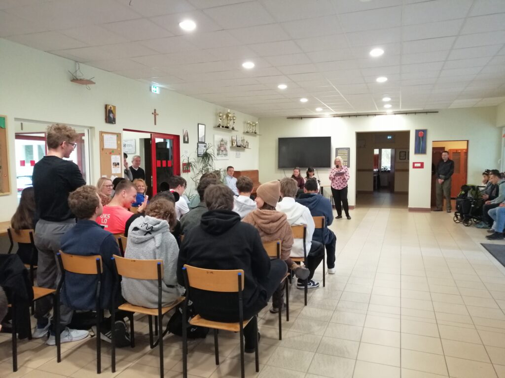 Zdjęcie ukazuje uczniów zgromadzonych na szkolnym korytarzu, siedzących w rzędach i zwróconych w kierunku głębi pomieszczenia. Wychowawczyni przemawia do uczniów wyczytując wychowanków swojej klasy.