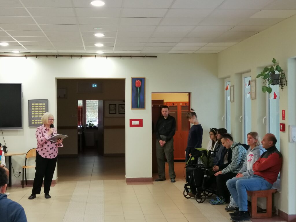 Zdjęcie ukazuje uczniów zgromadzonych na szkolnym korytarzu, siedzących w rzędach i zwróconych w kierunku głębi pomieszczenia. Wychowawczyni przemawia do uczniów wyczytując wychowanków swojej klasy.