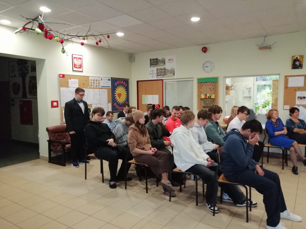 Zdjęcie ukazuje uczniów zgromadzonych na szkolnym korytarzu, siedzących w rzędach i zwróconych w kierunku głębi pomieszczenia. Wychowawczyni przemawia do uczniów wyczytując wychowanków swojej klasy.