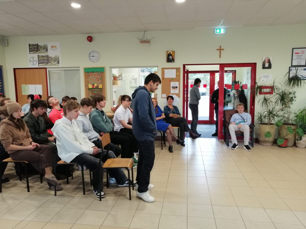 Zdjęcie ukazuje uczniów zgromadzonych na szkolnym korytarzu, siedzących w rzędach i zwróconych w kierunku głębi pomieszczenia. Wychowawczyni przemawia do uczniów wyczytując wychowanków swojej klasy.