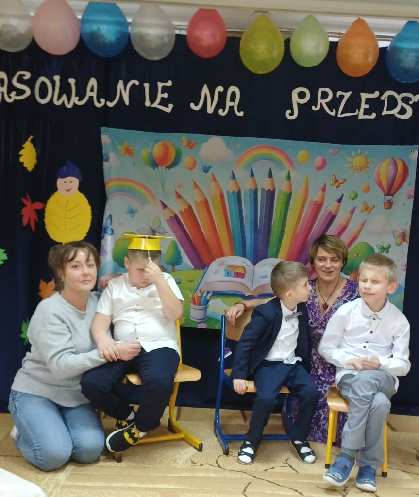 Elegancko ubrani chłopcy siedzą na krzesłach na tle dekoracji. Przy jednym klęczy mama, z tyłu wychowawczyni.