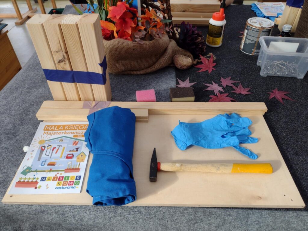 Na zdjęciu widoczny jest zestaw do majsterkowania, składający się z drewnianych elementów, narzędzi takich jak młotek i rękawiczki, oraz broszury "Mała Księga Majsterkowicz", ułożony na stole dekorowanym jesiennymi liśćmi.