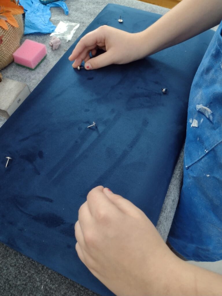 Na zdjęciu uczennica podnosi pinezki na stole z rozłożonym materiałem.