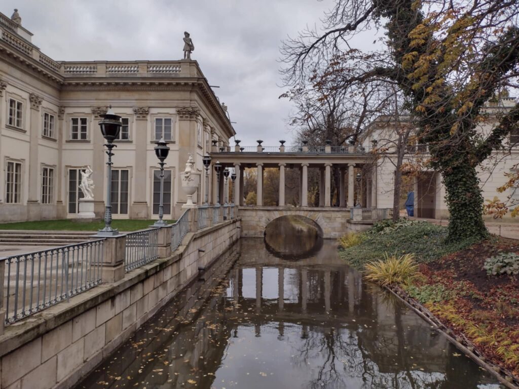     Zapytanie zostało przetworzone

Zdjęcie przedstawia klasycystyczny budynek, prawdopodobnie Pałac na Wyspie w Łazienkach Królewskich w Warszawie, otoczony kanałem wodnym z widocznym w głębi arkadowym mostem, co ma miejsce jesienią, o czym świadczą częściowo przebarwione liście na drzewach i na wodzie, pod szarym, pochmurnym niebem.