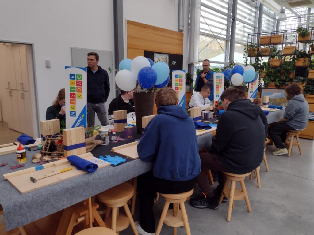 Przy stole, prawdopodobnie podczas warsztatów majsterkowania, siedzi grupa osób skupionych na pracy z drewnianymi elementami i narzędziami. W tle widać balony i rośliny, co tworzy atmosferę twórczego spotkania.