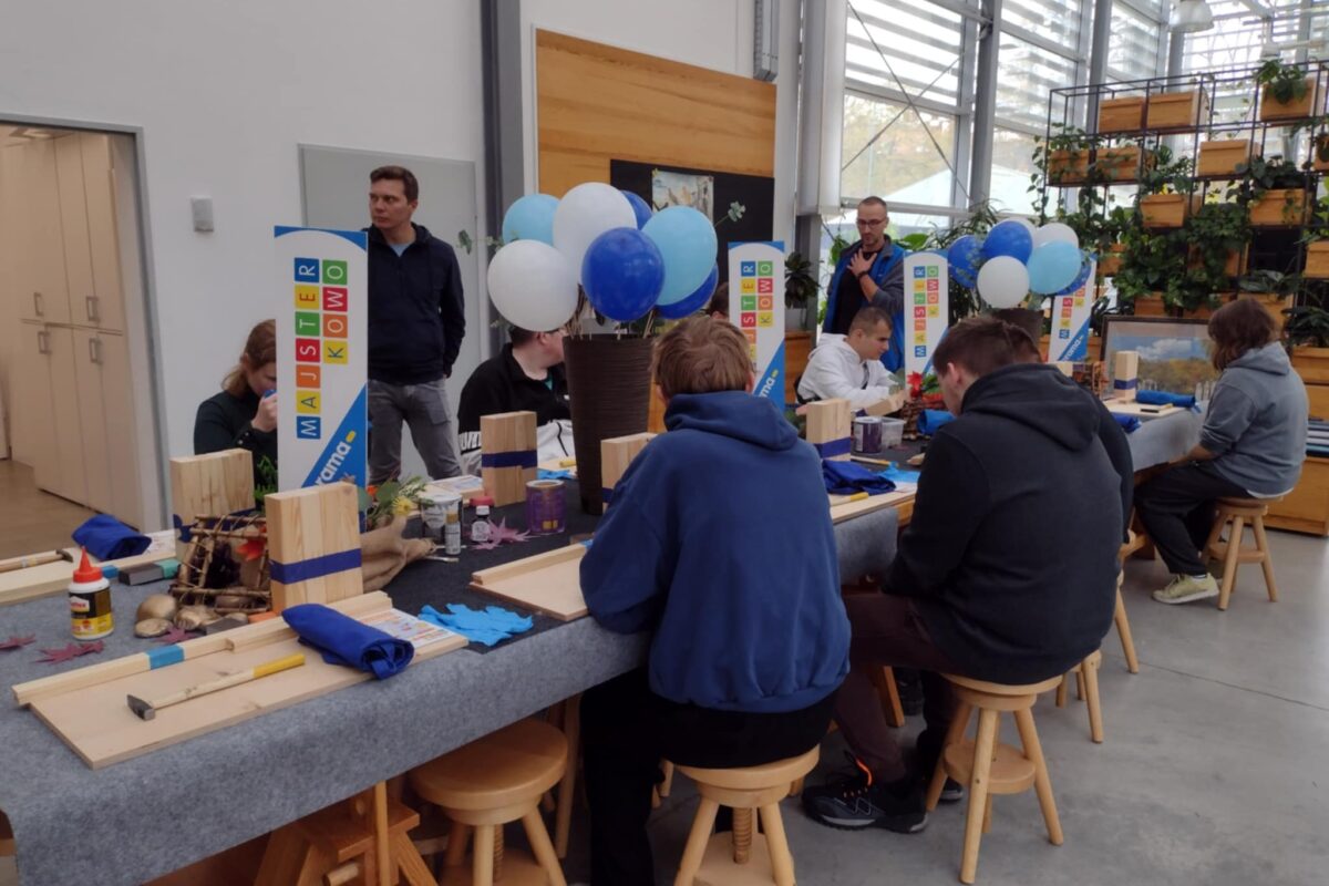 Przy stole, prawdopodobnie podczas warsztatów majsterkowania, siedzi grupa osób skupionych na pracy z drewnianymi elementami i narzędziami. W tle widać balony i rośliny, co tworzy atmosferę twórczego spotkania.
