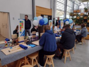 Przy stole, prawdopodobnie podczas warsztatów majsterkowania, siedzi grupa osób skupionych na pracy z drewnianymi elementami i narzędziami. W tle widać balony i rośliny, co tworzy atmosferę twórczego spotkania.