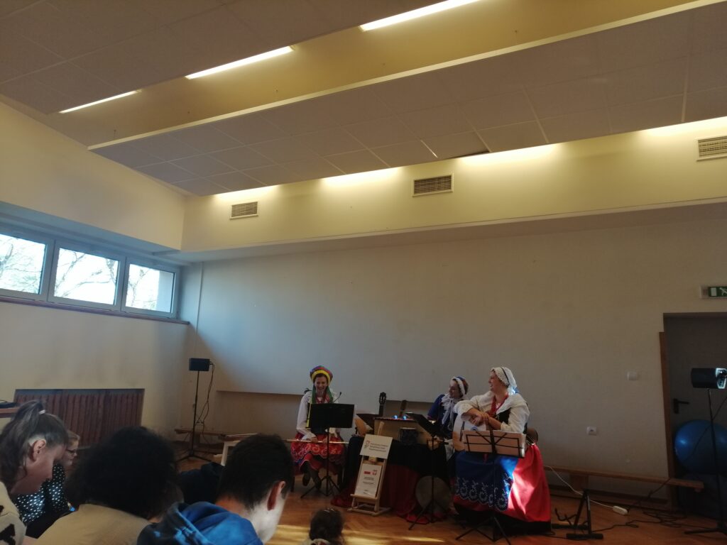 W dużej, jasnej sali trzy artystki z Fundacji „Szafa Kultury”, ubrane w regionalne stroje kujawskie, siedzą na scenie podczas interaktywnego spektaklu „W cieniu Mysiej Wieży”. Publiczność złożona z uczniów uważnie śledzi występ, który jest wzbogacony o wykorzystanie tradycyjnych instrumentów muzycznych.