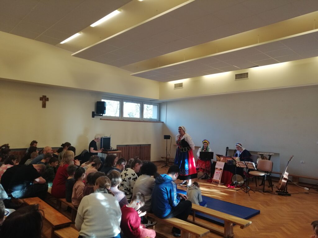 Duża widownia złożona z dzieci i dorosłych siedzi w rzędach, uważnie obserwując dwie artystki w tradycyjnych, regionalnych strojach, które prowadzą występ na niewielkiej scenie. Scena przedstawia moment aktywnej prezentacji w jasnej sali z oknami Panie prezentują przedstawienie.