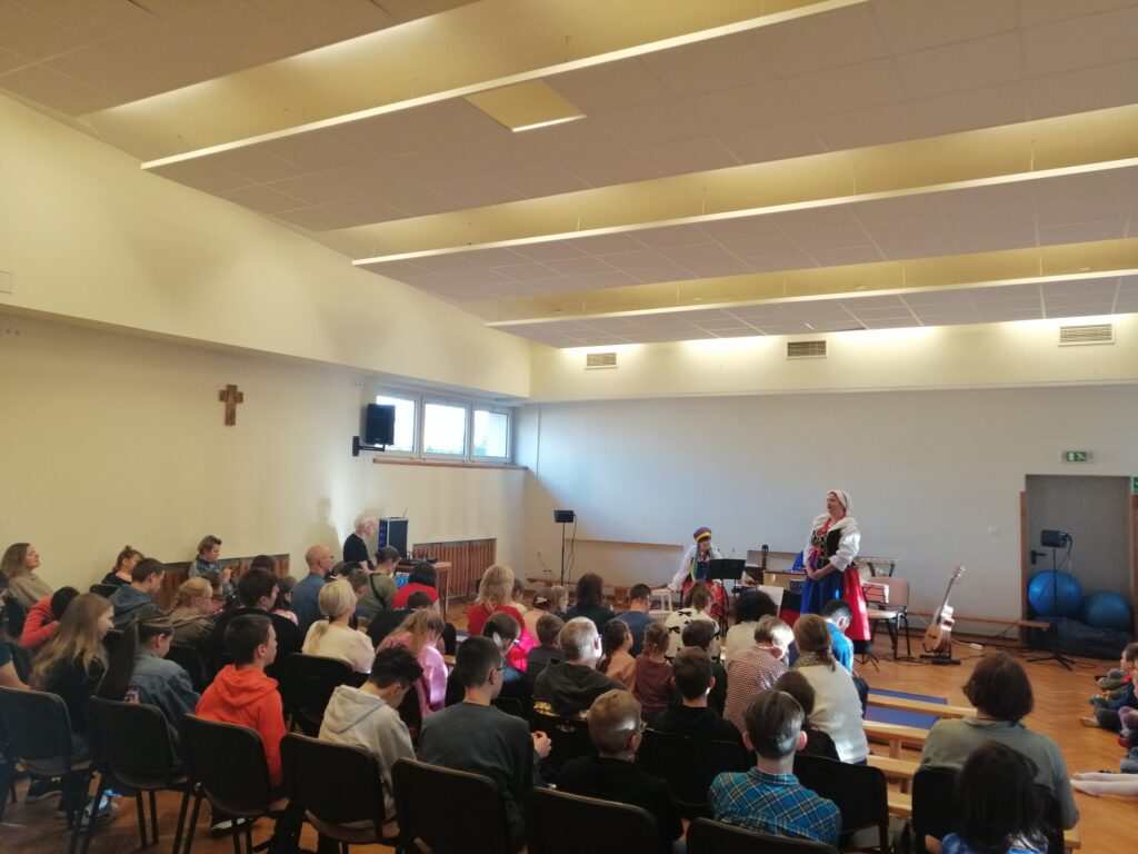 W dużej, jasnej sali szkolnej widzimy liczną publiczność, złożoną z dzieci i dorosłych, siedzącą w rzędach krzeseł, zwróconą w stronę sceny. Na niewielkim podwyższeniu dwie artystki w kolorowych, regionalnych strojach ludowych prowadzą występ, a jedna z nich aktywnie przemawia do widowni.