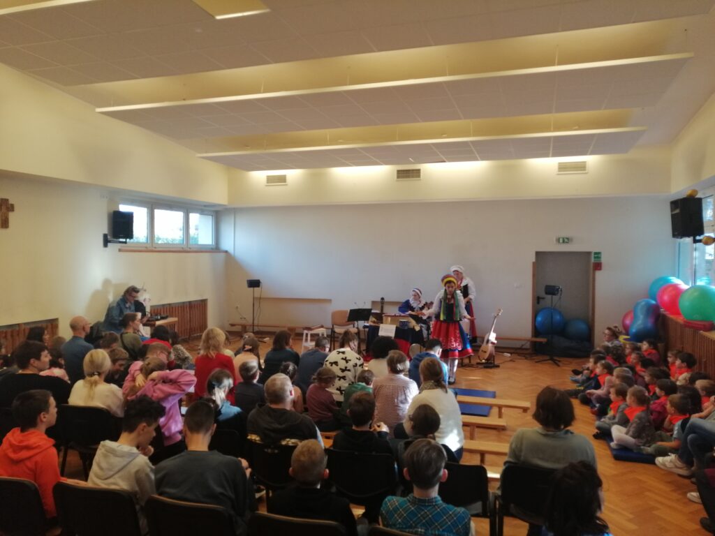 W dużej, jasnej sali szkolnej widzimy liczną publiczność, złożoną z dzieci i dorosłych, siedzącą w rzędach krzeseł, zwróconą w stronę sceny. Na niewielkim podwyższeniu dwie artystki w kolorowych, regionalnych strojach ludowych prowadzą występ, a jedna z nich aktywnie przemawia do widowni. Obok też siedzą uczniowie z innej szkoły zaproszone na występ