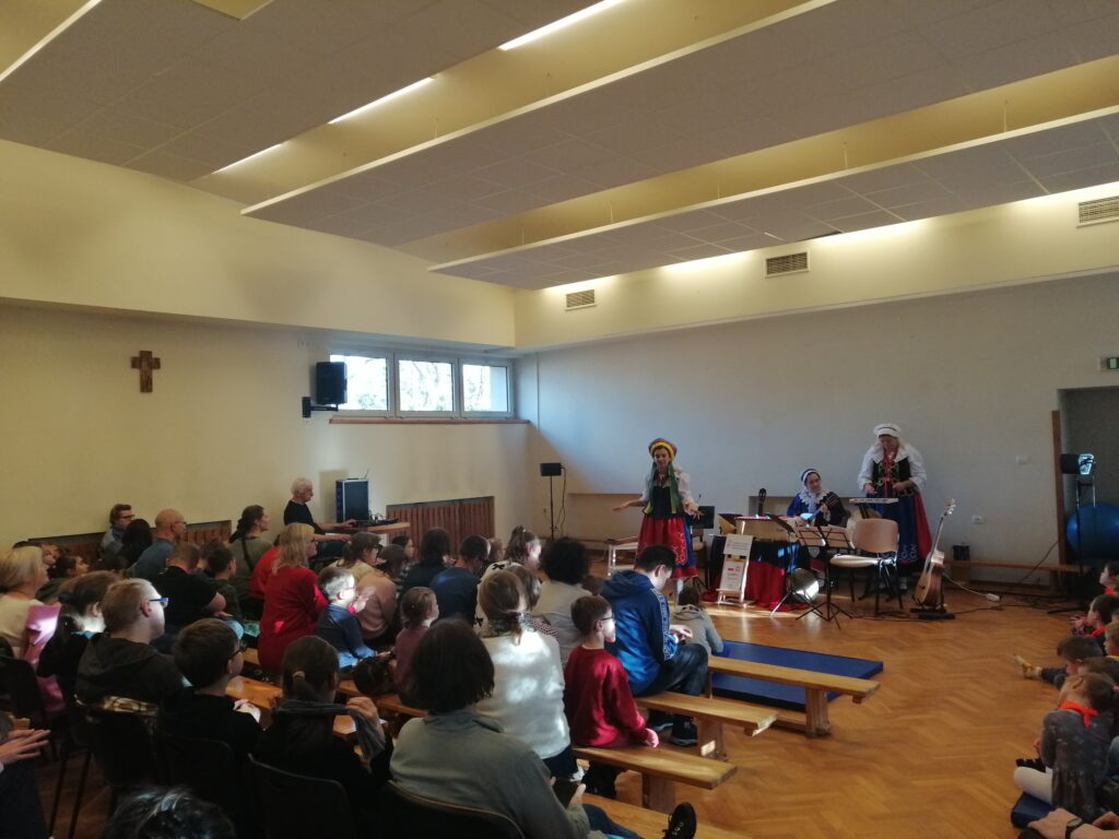 Trzy Panie Bajerki w kolorowych, tradycyjnych strojach ludowych aktywnie prowadzą występ na środku sali, przemawiając do licznej publiczności złożonej z dzieci i dorosłych. Widzowie siedzą na krzesłach, ławkach i podłodze, z zainteresowaniem obserwując dynamiczną prezentację folklorystyczną.