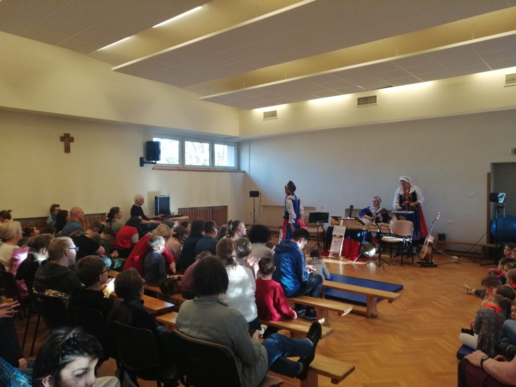 W dużej, jasnej sali z drewnianą podłogą, liczna publiczność, składająca się z dzieci i dorosłych, siedzi w rzędach krzeseł i na ławkach, oglądając występ. Na scenie trzy artystki w tradycyjnych strojach ludowych prowadzą prezentację, otoczone instrumentami muzycznymi.