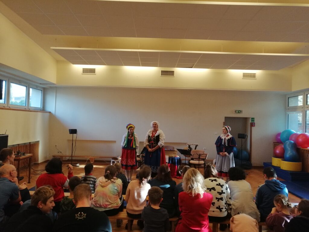 W dużej, jasnej sali z drewnianą podłogą, liczna publiczność, składająca się z dzieci i dorosłych, siedzi w rzędach krzeseł i na ławkach, oglądając występ. Na scenie trzy artystki w tradycyjnych strojach ludowych prowadzą prezentację, otoczone instrumentami muzycznymi.