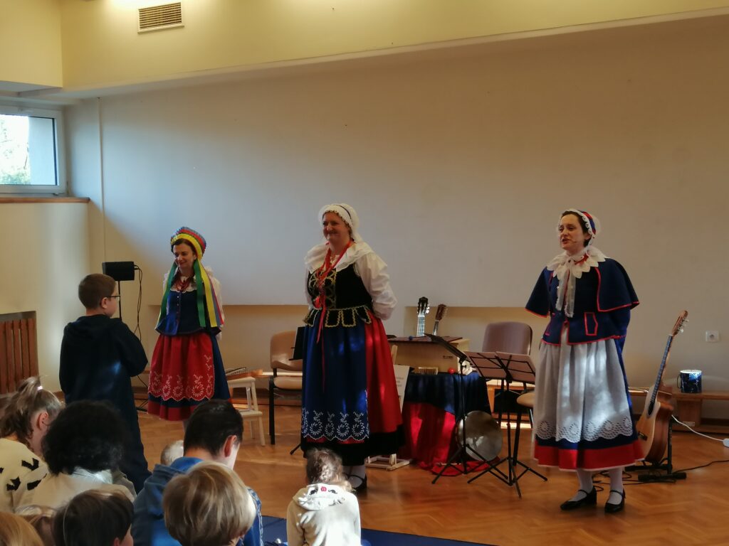 Panie w kolorowych, tradycyjnych strojach ludowych stoją na środku sali,  dziękując publiczności za uwagę po zakończonym występie. Wokół nich siedzi widownia, składająca się z dzieci i dorosłych.