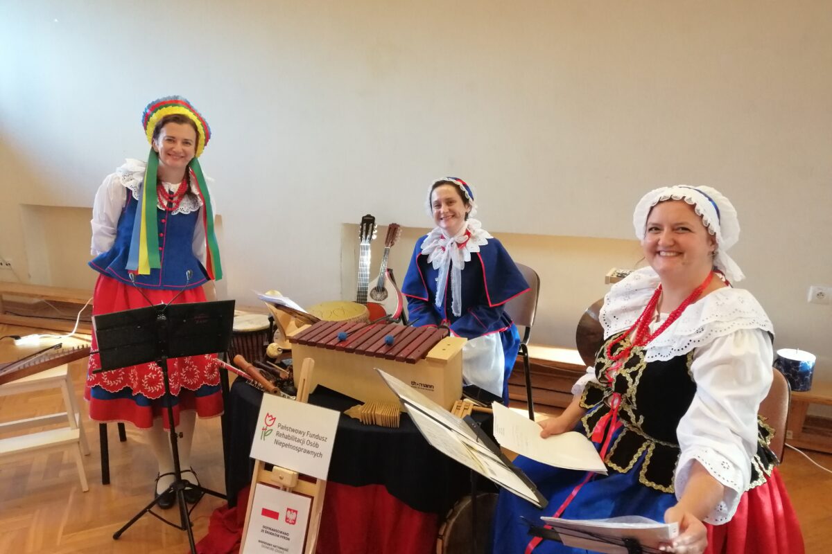 Trzy uśmiechnięte kobiety w tradycyjnych, kolorowych polskich strojach ludowych stoją i siedzą za stołem, na którym prezentują bogatą kolekcję ludowych instrumentów muzycznych, w tym duży ksylofon. Scena ma charakter kulturalnej prezentacji i emanuje pozytywną energią oraz radością.