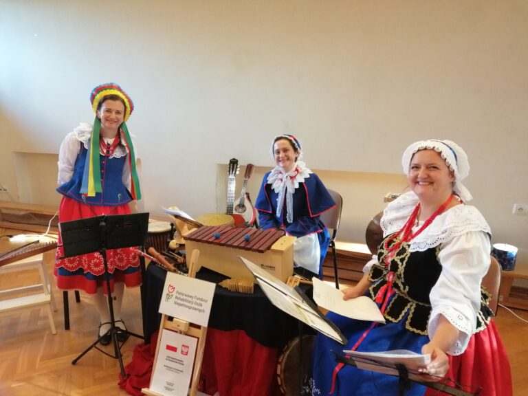 Trzy uśmiechnięte kobiety w tradycyjnych, kolorowych polskich strojach ludowych stoją i siedzą za stołem, na którym prezentują bogatą kolekcję ludowych instrumentów muzycznych, w tym duży ksylofon. Scena ma charakter kulturalnej prezentacji i emanuje pozytywną energią oraz radością.