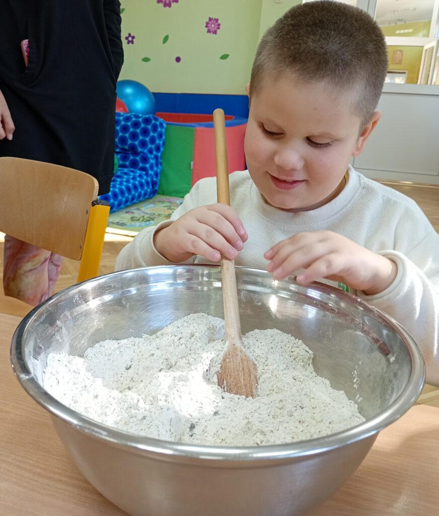 Chłopiec siedzi przy stole, miesza w misce  drewnianą łyżką składniki ciasta.