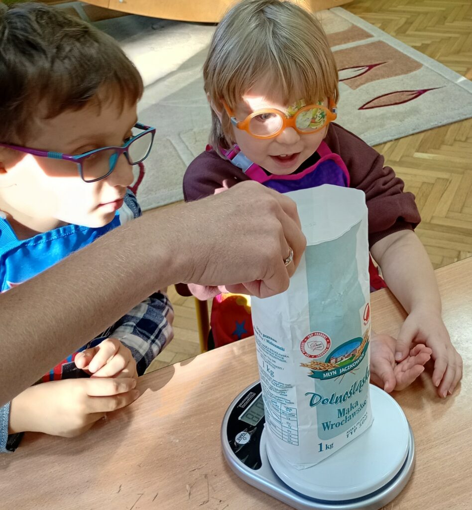 Chłopiec i dziewczynka siedzą przy stole, przed nimi na wadze stoi mąka. 