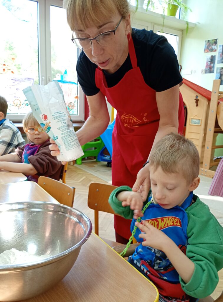 Chłopiec siedzi przy stole, przed nim miska z mąką, obok stoi nauczycielka z opakowaniem mąki w ręce.