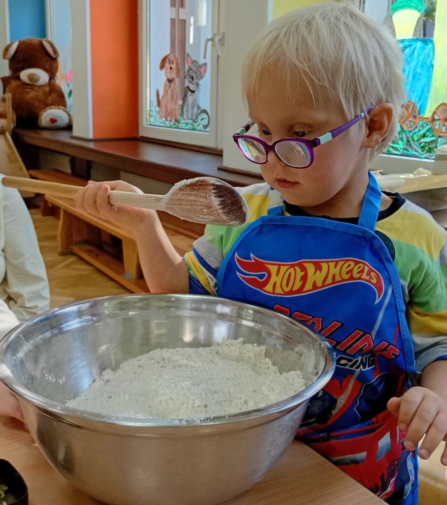 Chłopiec siedzi przy stole, przed nim miska z mąką, w ręce trzyma drewnianą łyżkę.