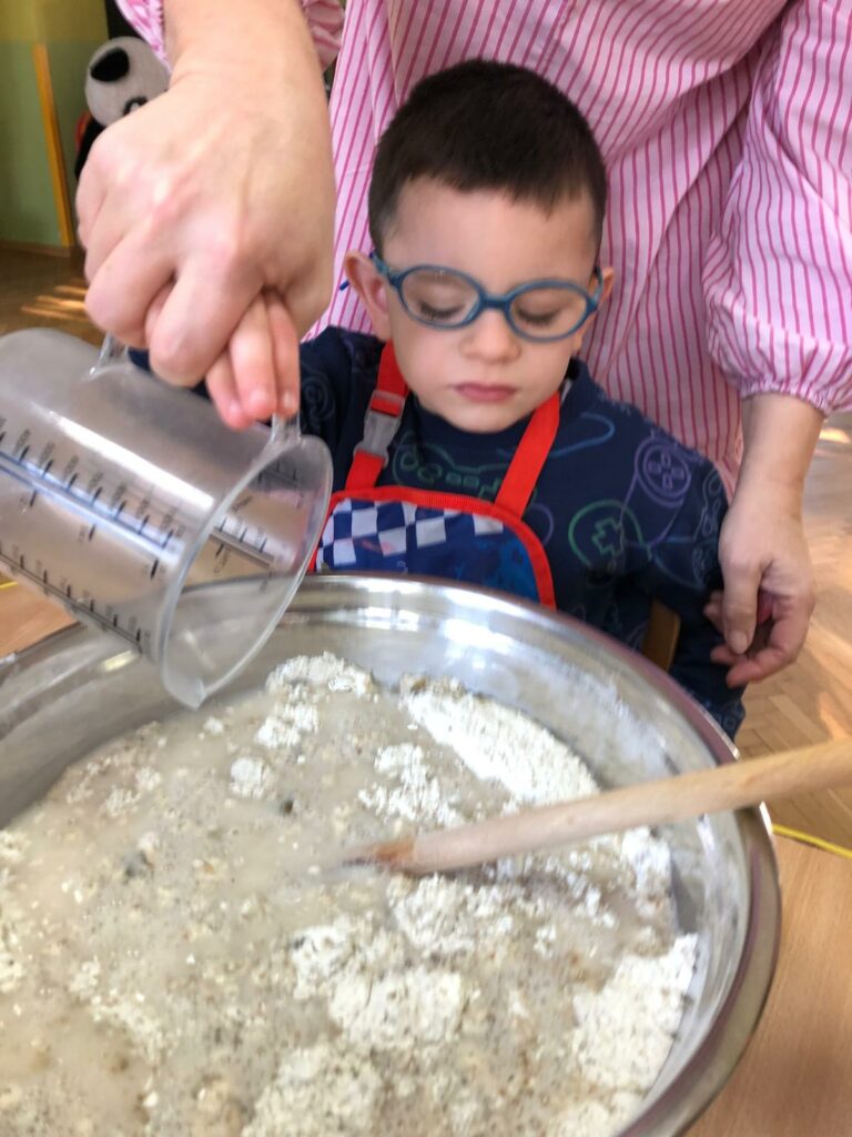 Chłopiec siedzi przy stole, przed nim stoi miska z ciastem na chleb, dolewa wody do ciasta.