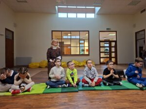 Na zdjęciu widać grupę najmłodszych uczniów, którzy siedzą na matach na podłodze, czekając na swoją kolej podczas Konkursu Recytatorskiego dla klas 1-3 o tematyce zwierzęcej. W tle, na środku kadru, stoi uśmiechnięta nauczycielka, a całe wydarzenie odbywało się w przyjaznej, pełnej entuzjazmu atmosferze.