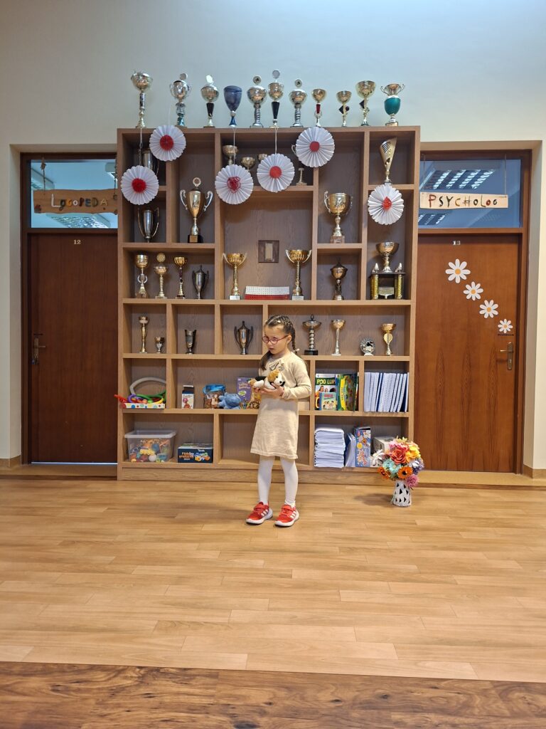 Na zdjęciu widzimy uczennice, ubraną w jasną sukienkę, stojącą w szkolnym korytarzu przed dużą drewnianą gablotą wypełnioną pucharami i ozdobioną białymi wachlarzami. Dziewczynka, trzyma w ręku pieska pluszowego, a za nią widoczne są drzwi do gabinetów Logopedy i Psychologa.