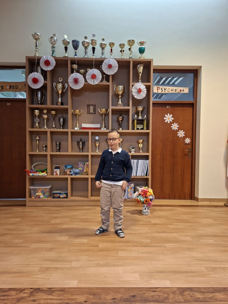 Chłopiec w okularach stojący na tle gabloty z pucharami w niebieskim swetrze.