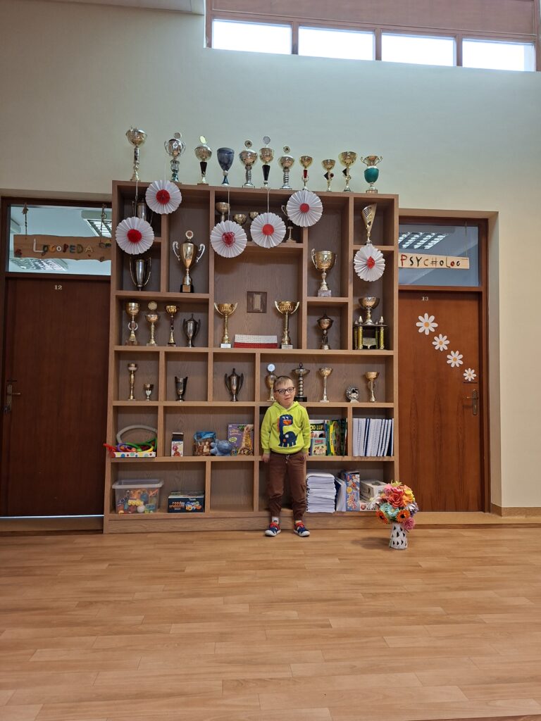 Uśmiechnięty chłopiec stojący na tle gabloty z pucharami w jaskrawozielonej bluzie, w szkolnym korytarzu.