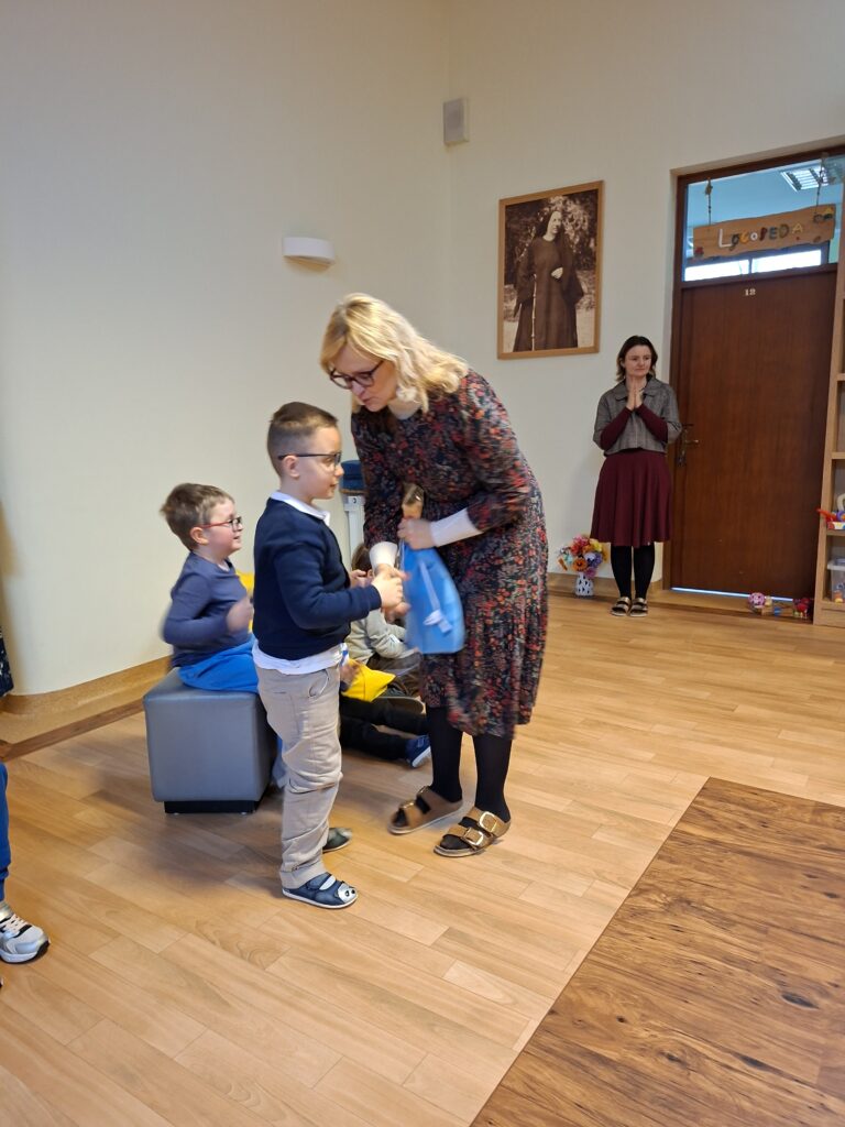 Na zdjęciu widzimy ucznia, który  odbiera nagrodę w Konkursie Recytatorskim dla klas 1-3 o tematyce zwierzęcej, wręczaną mu przez jurorkę w obecności innych uczniów. Chłopiec trzyma niebieski worek z nagrodą.