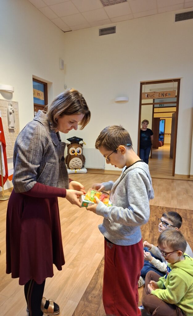 Na zdjęciu widzimy chłopca odbierającego nagrodę w postaci kolorowego pudełka z rąk jurorki lub organizatorki. Ten moment nagradzania jest częścią finału Konkursu Recytatorskiego dla klas 1-3, który przebiegał w przyjaznej, pełnej entuzjazmu atmosferze