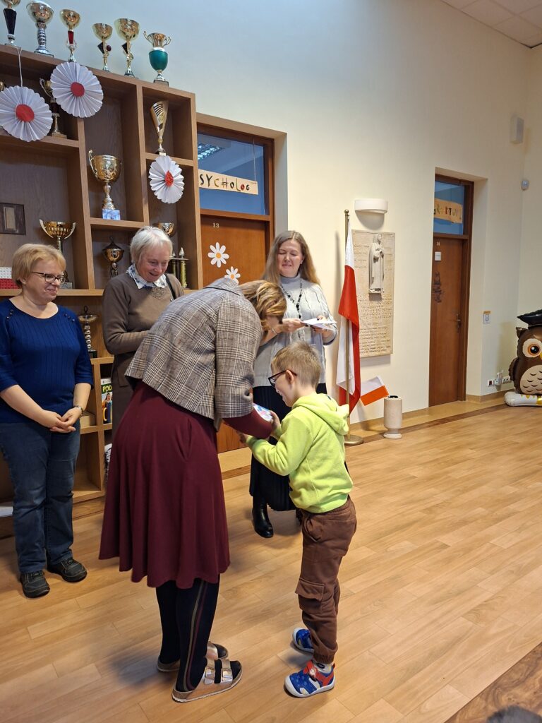 Uczeń odbiera nagrodę z rąk jurorki podczas finału Konkursu Recytatorskiego dla klas 1-3. W tle widać uśmiechnięte Panie i szafkę z pucharami.