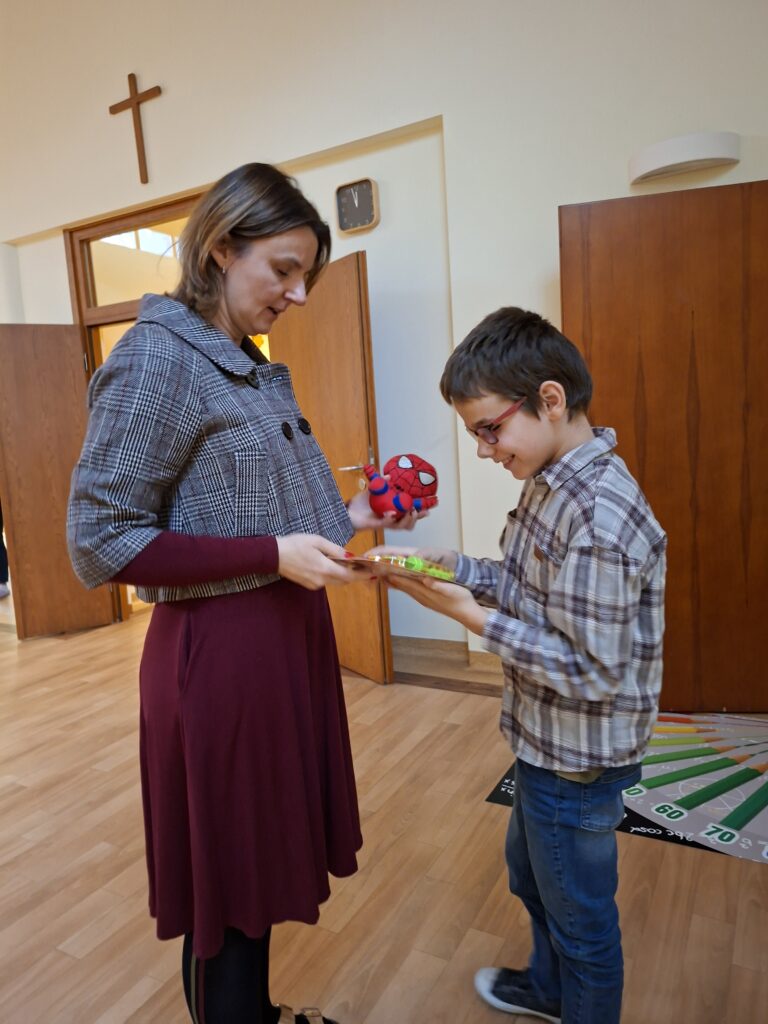Na zdjęciu widzimy uśmiechniętego ucznia odbierającego nagrodę w postaci spider mana i zabawki w pudełku. Ten moment nagradzania jest częścią finału Konkursu Recytatorskiego.