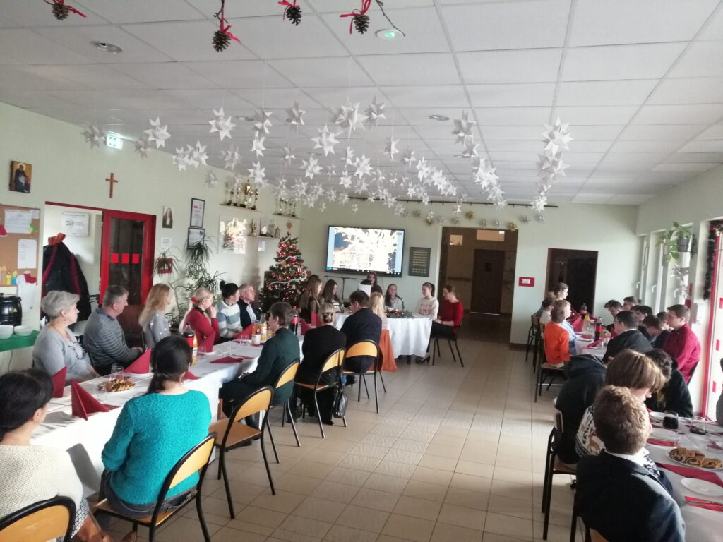 Zdjęcie przedstawia społeczność Jabłonek – uczniów i nauczycieli – celebrujących spotkanie przedświąteczne przy wspólnym stole w udekorowanym holu szkoły, podczas którego odbywają się występy artystyczne oraz wręczanie nagród konkursowych.