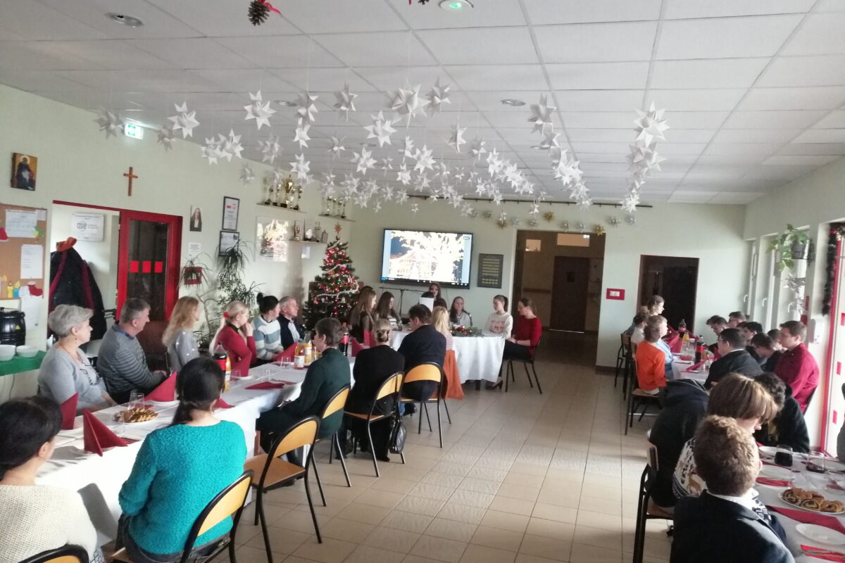 Zdjęcie przedstawia społeczność Jabłonek – uczniów i nauczycieli – celebrujących spotkanie przedświąteczne przy wspólnym stole w udekorowanym holu szkoły, podczas którego odbywają się występy artystyczne oraz wręczanie nagród konkursowych.