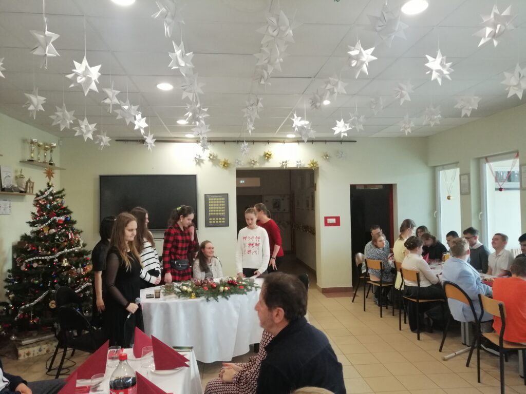 Zdjęcie przedstawia uczennice klas III i IV Liceum oraz IV Technikum, Stoją obok choinki i wystrojonego stołu stojąc w trakcie przedstawienia.