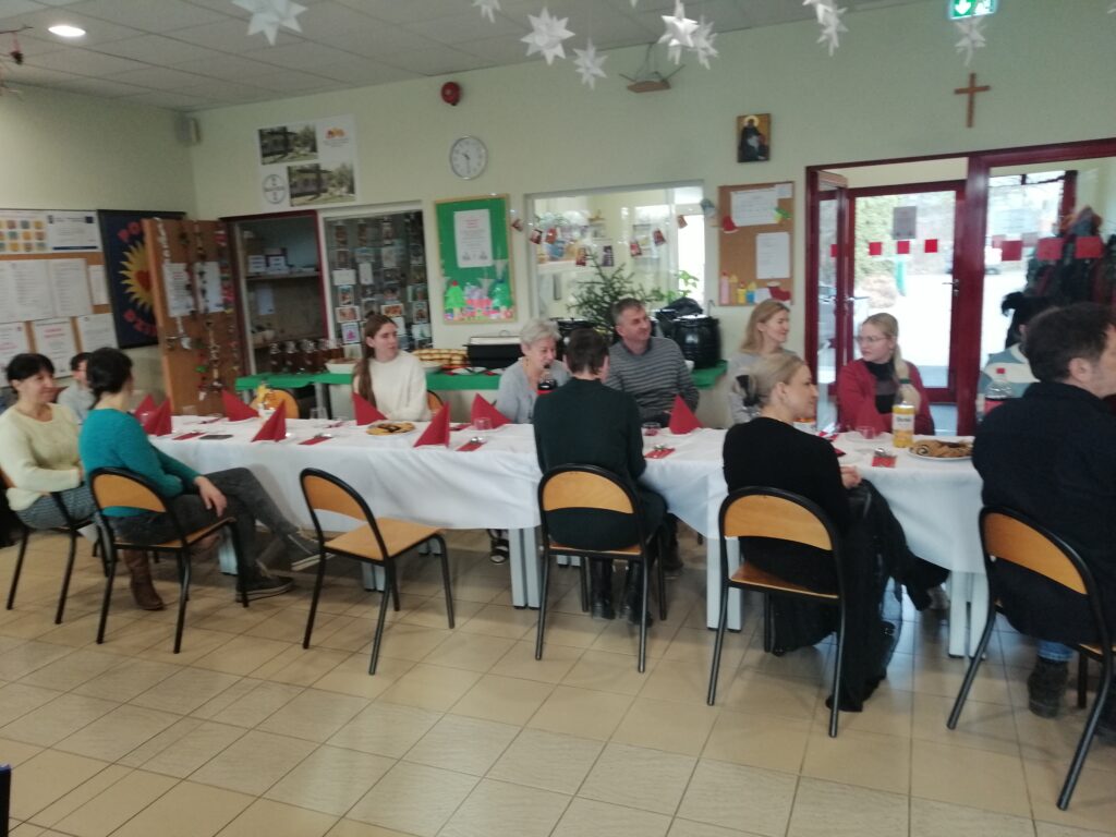 Uczestnicy spotkania słuchają piosenki siedząc  przy stole. W tle widać wystrój kiermaszu świątecznego.