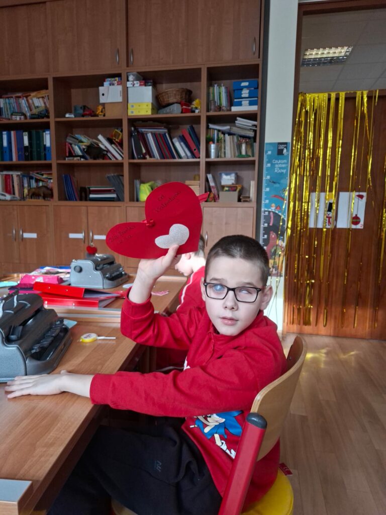 Uczeń ubrany w czerwoną bluzę siedzi przy biurku z maszyną brajlowską i unosi w górę duże, czerwone serce z dedykacją, przygotowane w ramach poczty walentynkowej.