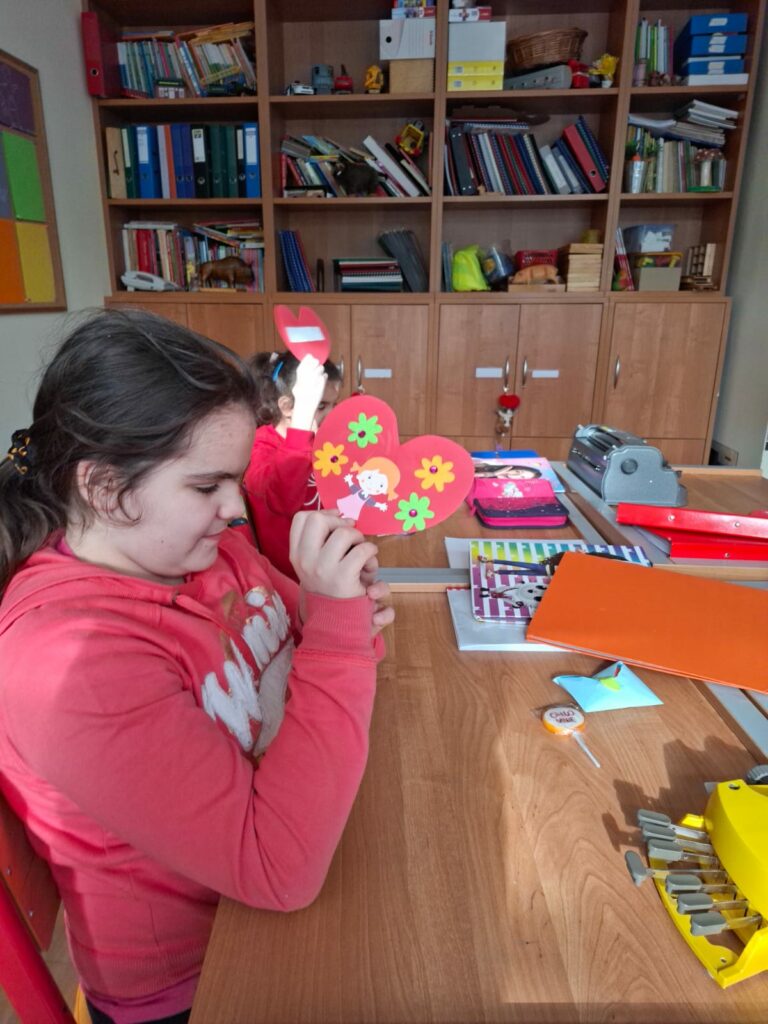 Dziewczyny w klasie trzymają w dłoniach duże, czerwone serca ozdobione kwiatkami siedząc przy szkolnej ławce.