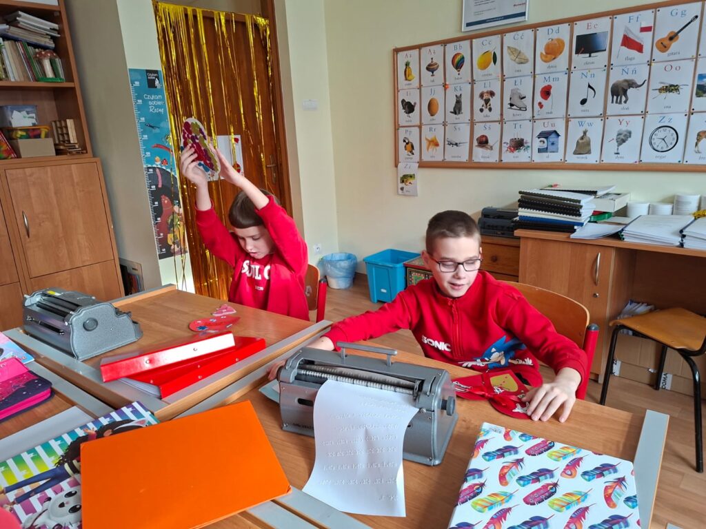 Dwóch uczniów siedzi przy ławkach z maszynami brajlowskimi, przygotowując i prezentując kolorowe, zdobione serca.