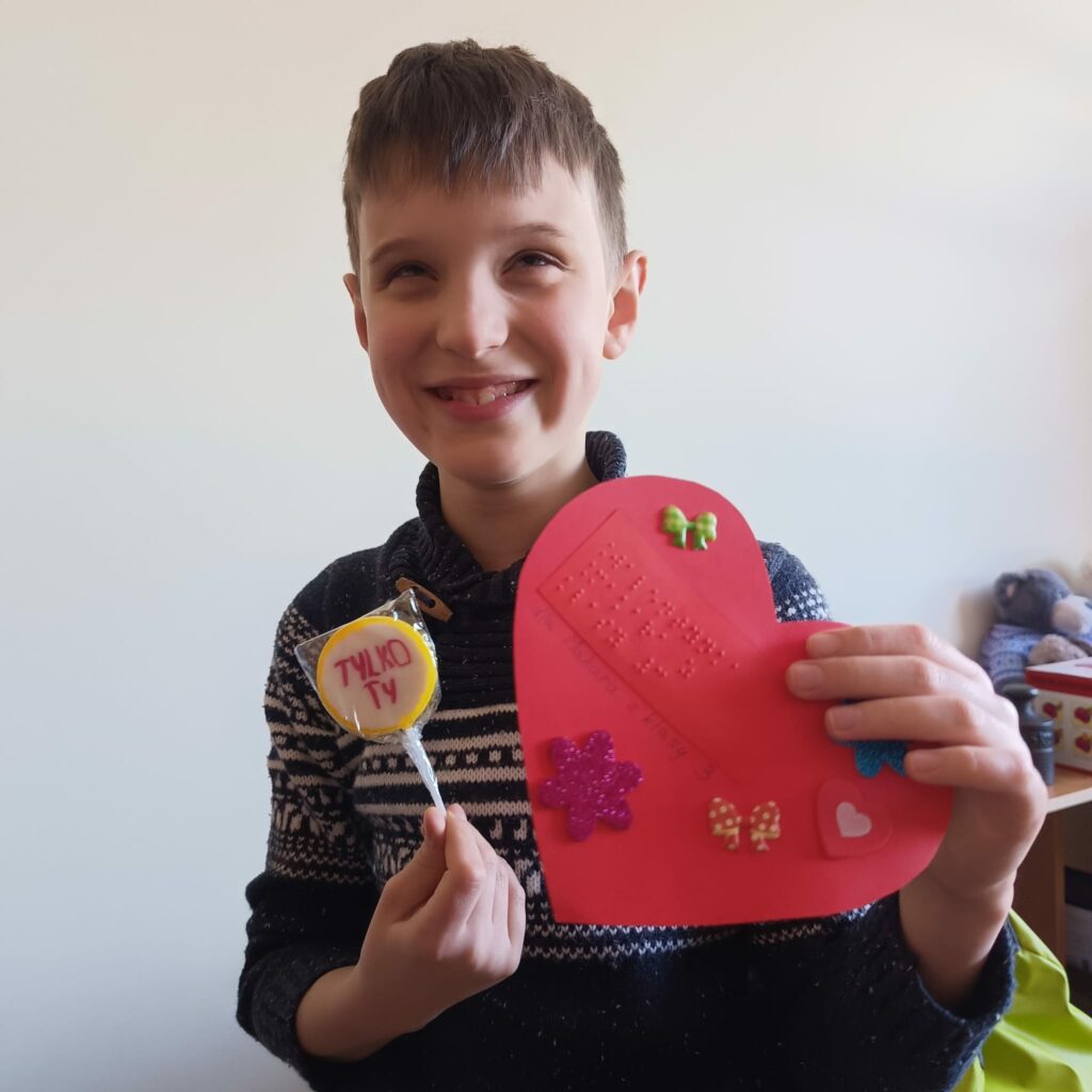 Uczeń trzyma czerwone serce z napisem brajlowskim oraz lizaka z napisem „TYLKO TY”.