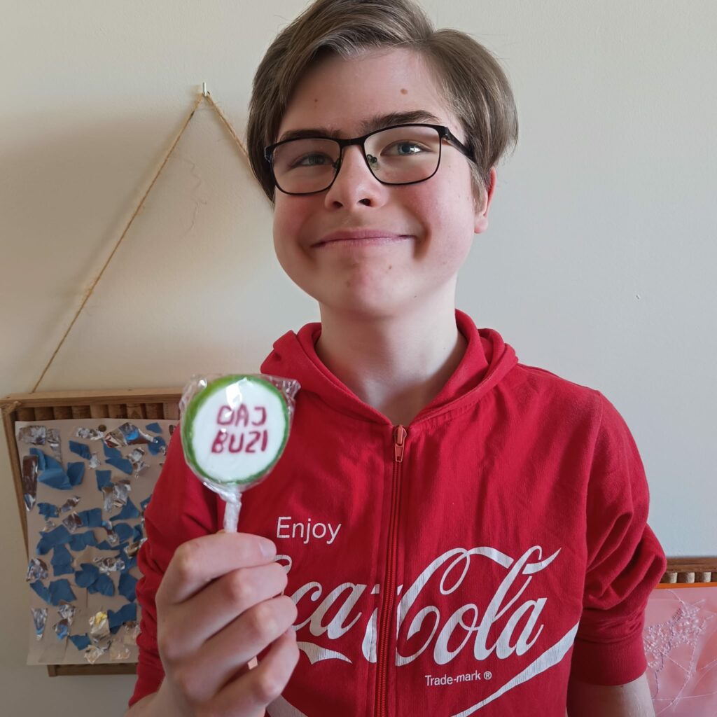 Uśmiechnięty uczeń w okularach i czerwonej bluzie prezentuje do aparatu okrągłego walentynkowego lizaka z napisem „DAJ BUZI”.